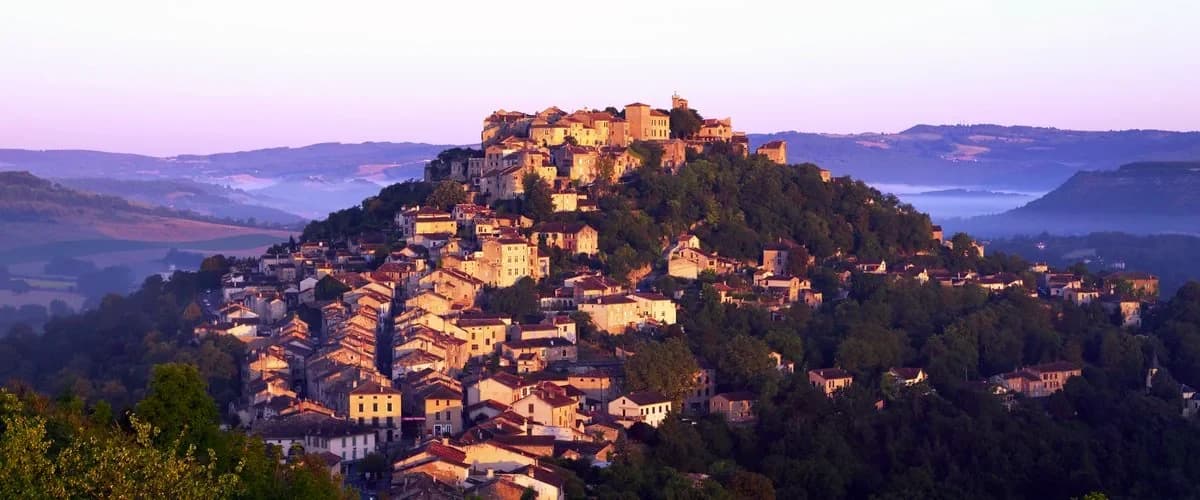 Village médiéval aux maisons de pierre blonde étagées sur une colline boisée, éclairé par le soleil levant, avec des vallées noyées dans la brume en contrebas